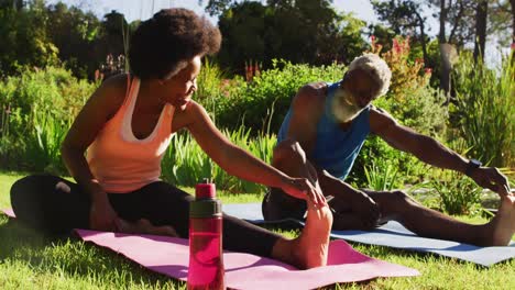 una pareja de ancianos afroamericanos haciendo ejercicio al aire libre sentados estirándose en un jardín soleado
