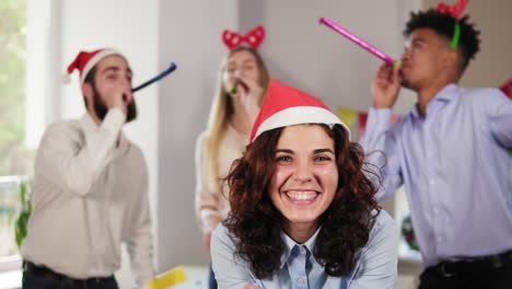 joven mujer atractiva soplando confeti dorado de las manos con sombrero de navidad mientras sus compañeros de trabajo de fiesta en el fondo