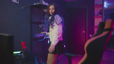 woman carrying popcorn walks toward ergonomic chair seats down adjusts posture under vibrant neon studio lighting wearing fluffy sweater and fishnet tights preparing for cozy relaxation session