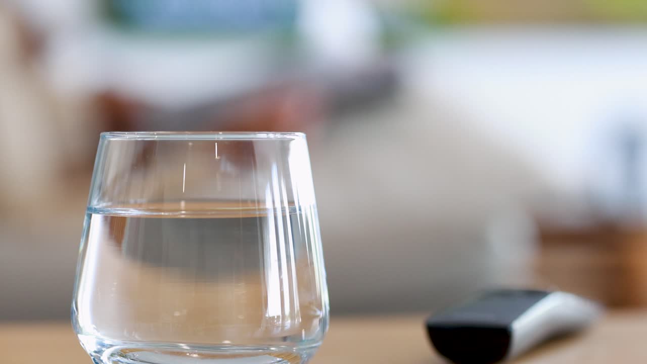 A glass of water and TV remote sit on a table in a bright living room as a woman reaches for the remote, background softly blurred