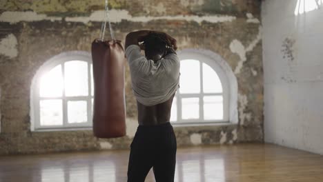 African-american-athlete-walking-into-room-with-boxing-pear,-taking-off-shirt