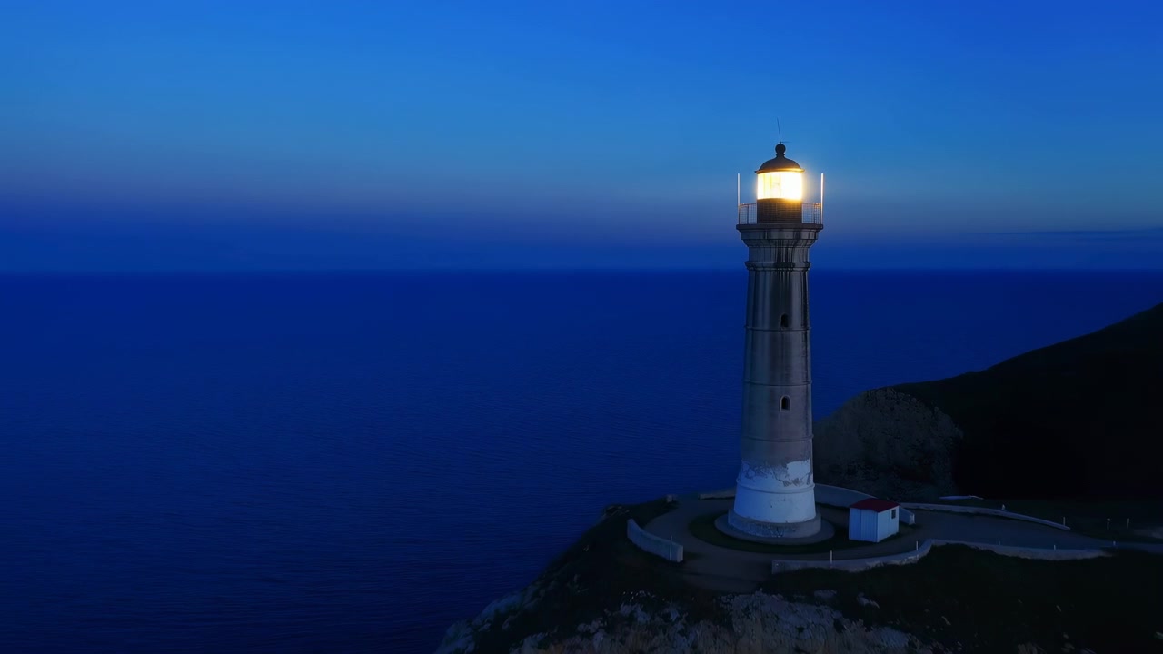 Composizione aerea mostra un faro luminoso sopra una costa frastagliata mentre la vista della telecamera si allontana, rivelando l'acqua circostante, le texture delle scogliere, il cielo blu intenso e un paesaggio costiero pacifico