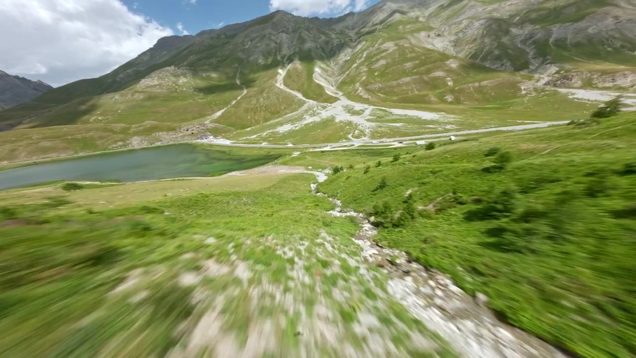 Drone FPV da corsa sfiora una luminosa collina alpina verde verso un piccolo lago e una strada in un'ampia valle italiana