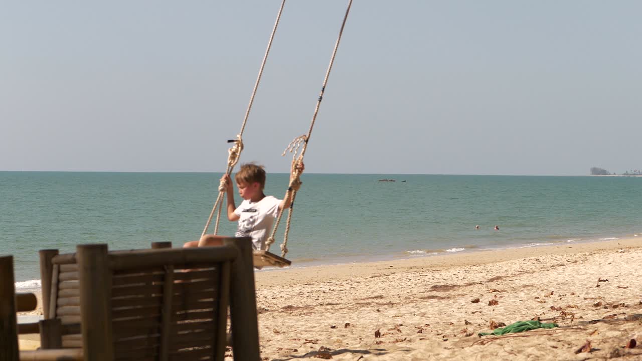 niño en un columpio, en la playa de pak weep, el mar de andaman en el fondo, día soleado, en khao lak, tailandia - cámara lenta