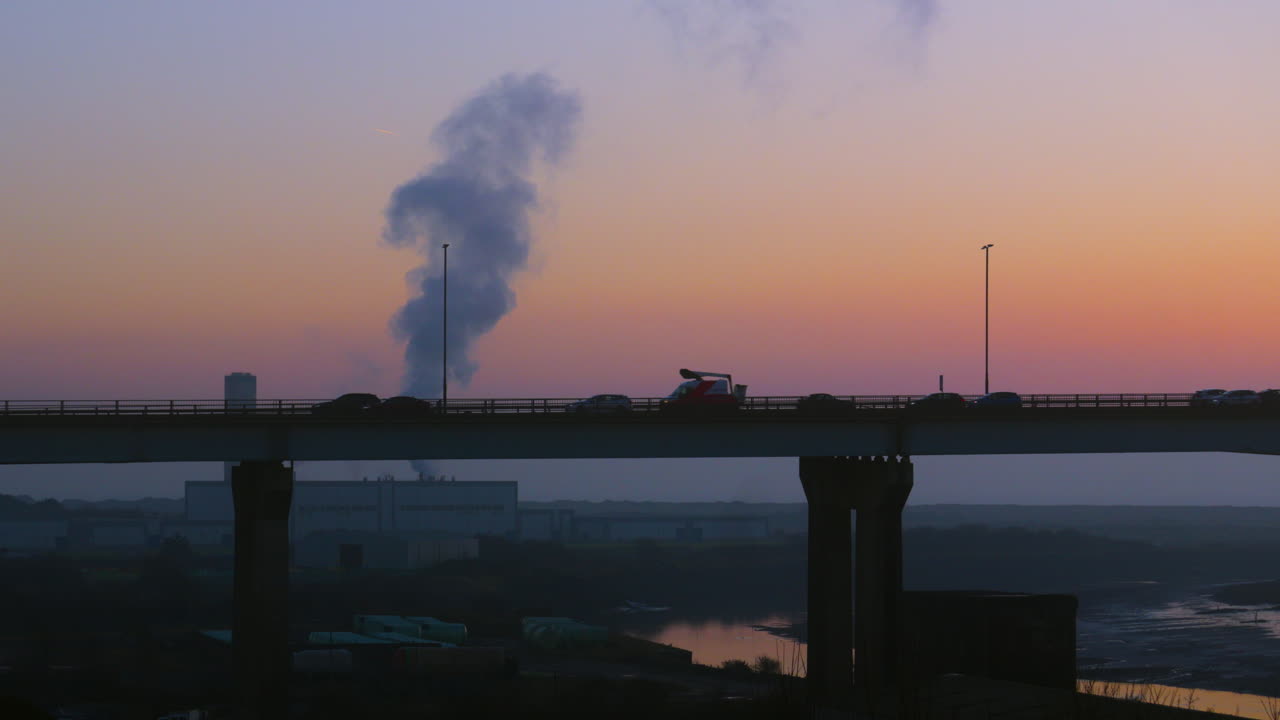 View of Cars and Trucks Crossing Bridge at Sunset with Factory Releasing Silhouetted Pollutants into Atmosphere in Background with River Nearby. Filmed on M4 Motorway Near Swansea, Wales 4K.