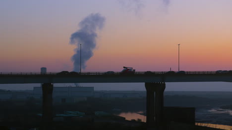 view of cars and trucks crossing bridge at sunset with factory releasing silhouetted pollutants into atmosphere in background with river nearby. filmed on m4 motorway near swansea, wales 4k.