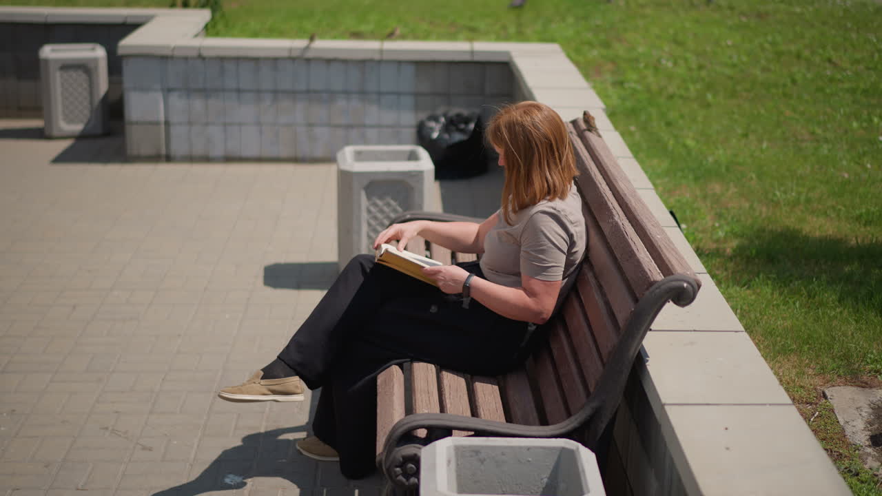 Woman sitting on wooden bench reading book outdoors on sunny day, small bird perched beside her adding peaceful and natural charm, green grass and trees behind