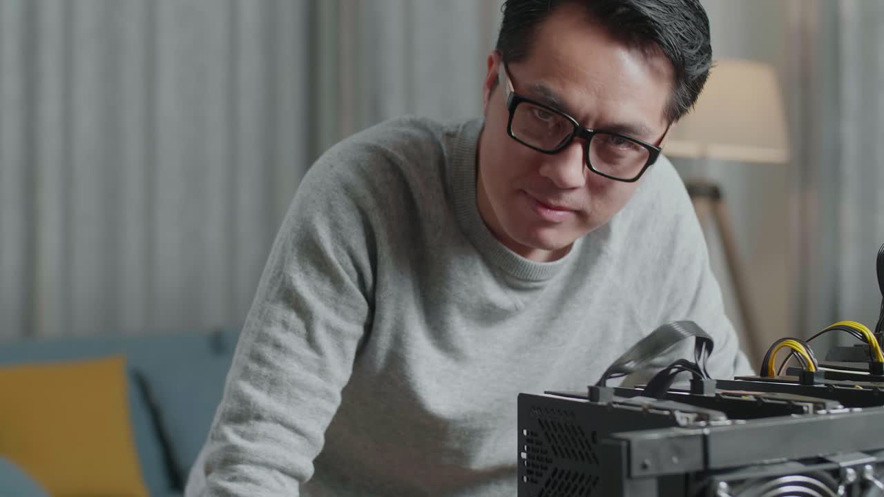 안경을 쓴 아시아 남자가 카메라에 미소 짓고 노트북과 광산 기계를 사용하여 암호화폐를 채굴합니다.