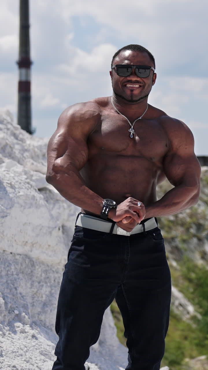 Shirtless afro american sportsman outdoors. Muscular bodybuilder with dark skin in sunglasses posing on hill on factory background. Vertical video