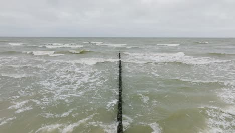 Establishing-aerial-view-of-Baltic-sea-coast-on-a-overcast-day,-old-wooden-pier,-white-sand-beach,-large-storm-waves-crushing-against-the-coast,-climate-changes,-wide-drone-shot-moving-forward-low