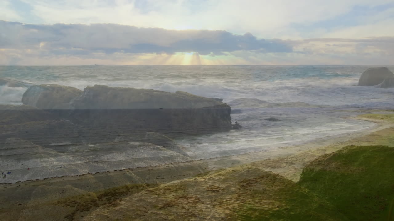 Crashing waves against rocky coastline under cloudy sky with sun rays filtering