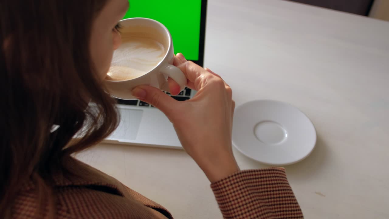 Back view - woman sitting at table drinking coffee and looking at laptop