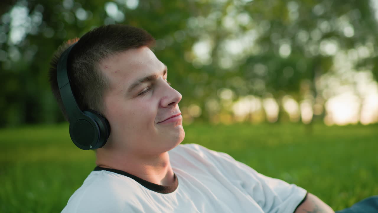 close up de artista de música con auriculares, asintiendo con la cabeza y disfrutando de la música con una sutil sonrisa mientras mira hacia arriba, el fondo presenta vegetación vibrante, árboles y luz del sol
