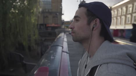 close view of a young man resting and enjoying the moment while listening to music with headphones the man is standing on a bridge above a river canal in the city of london, england