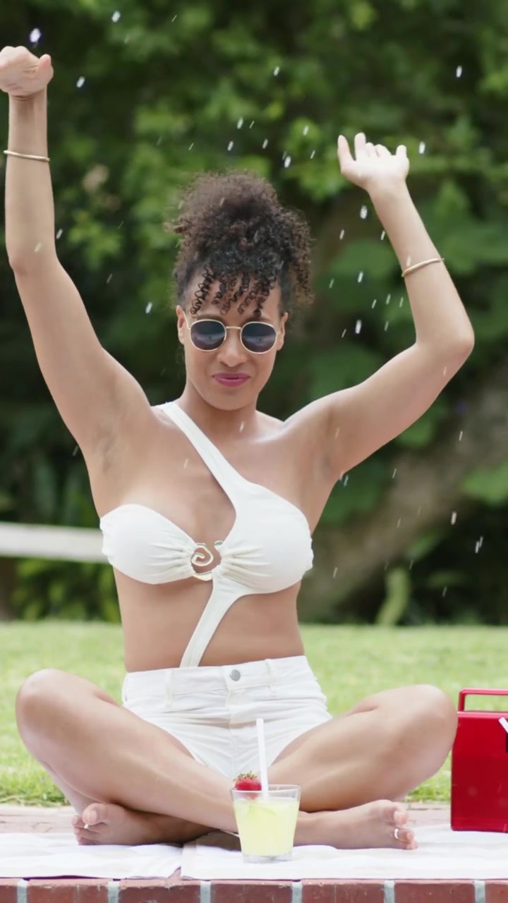 Donna in costume da bagno bianco che alza le braccia, reagendo a uno schizzo sul bordo della piscina, con bevanda e borsa frigo rossa