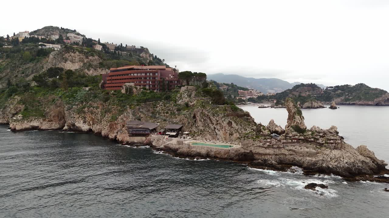 Aerial drone wide shot panning left of a fancy hotel perched on the cliff, overlooking the stunning views of Taormina, Sicily, Italy (Taormina, Sicilia, Italia)
