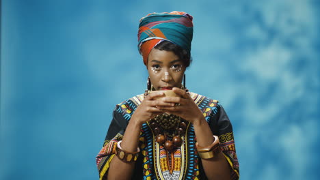 african american young beautiful woman in traditional clothes and turban drinking hot tea and looking at camera