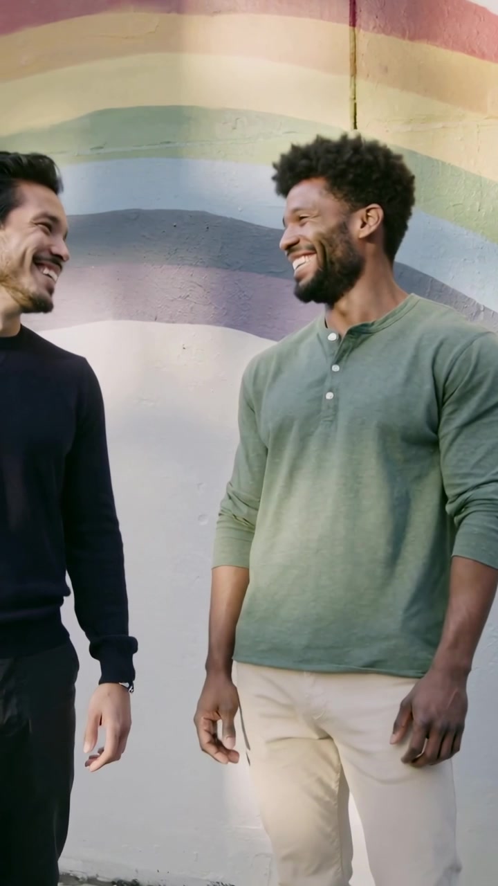 Video verticale: Sguardi che si incontrano, due uomini che condividono una battuta, sorridendo accanto a un murale arcobaleno, una maglietta henley verde visibile