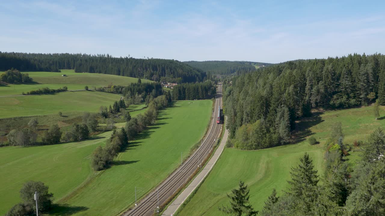 drone de un tren rojo avanzando en una línea ferroviaria de doble vía, cortando a través de un paisaje bucólico de campos y árboles, y acercándose a un túnel lejano bajo la claridad de un cielo azul