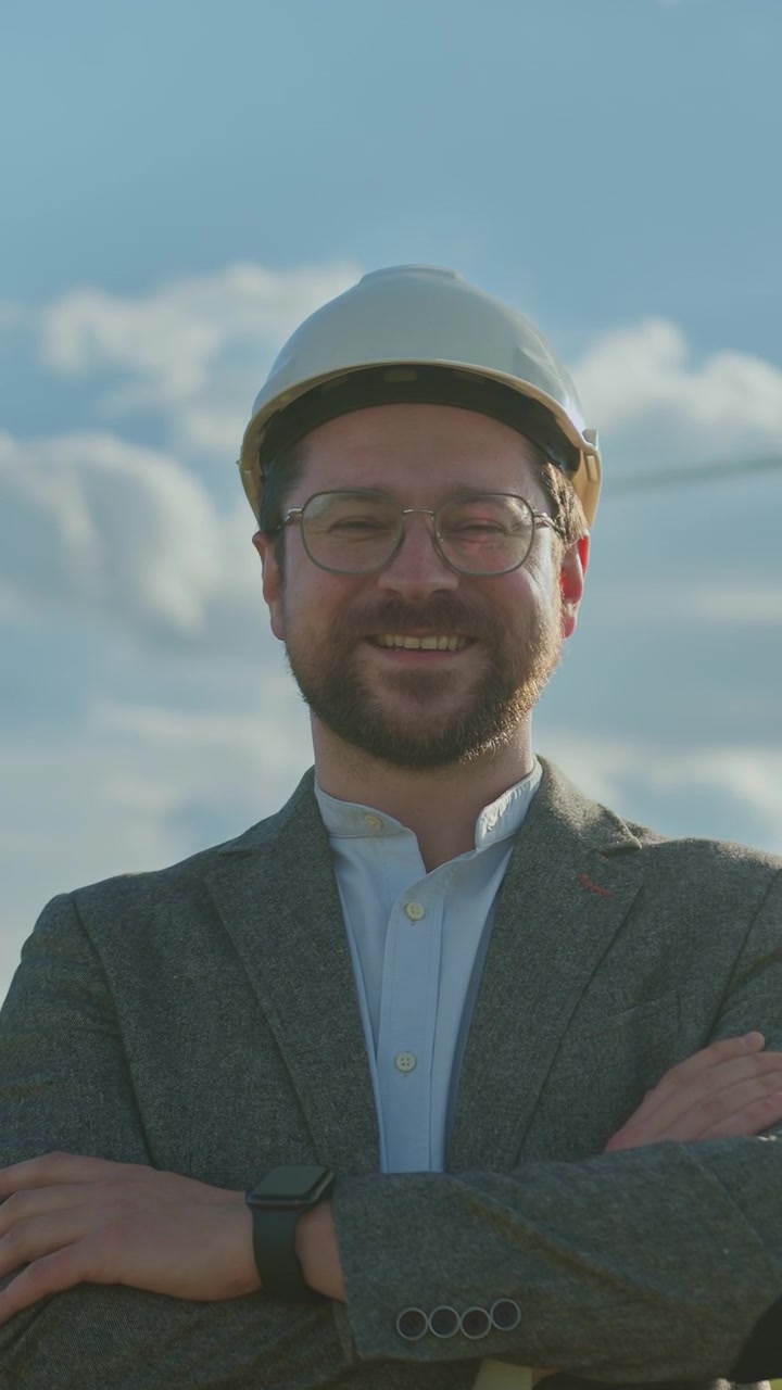 Hombre con sombrero y gafas, con los brazos cruzados, sonríe al aire libre cerca de aerogeneradores en un día nublado