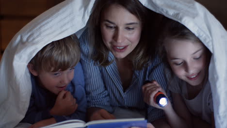 vista de cerca de un niño lindo y su hermana usando una linterna y leyendo un libro debajo de la manta por la noche