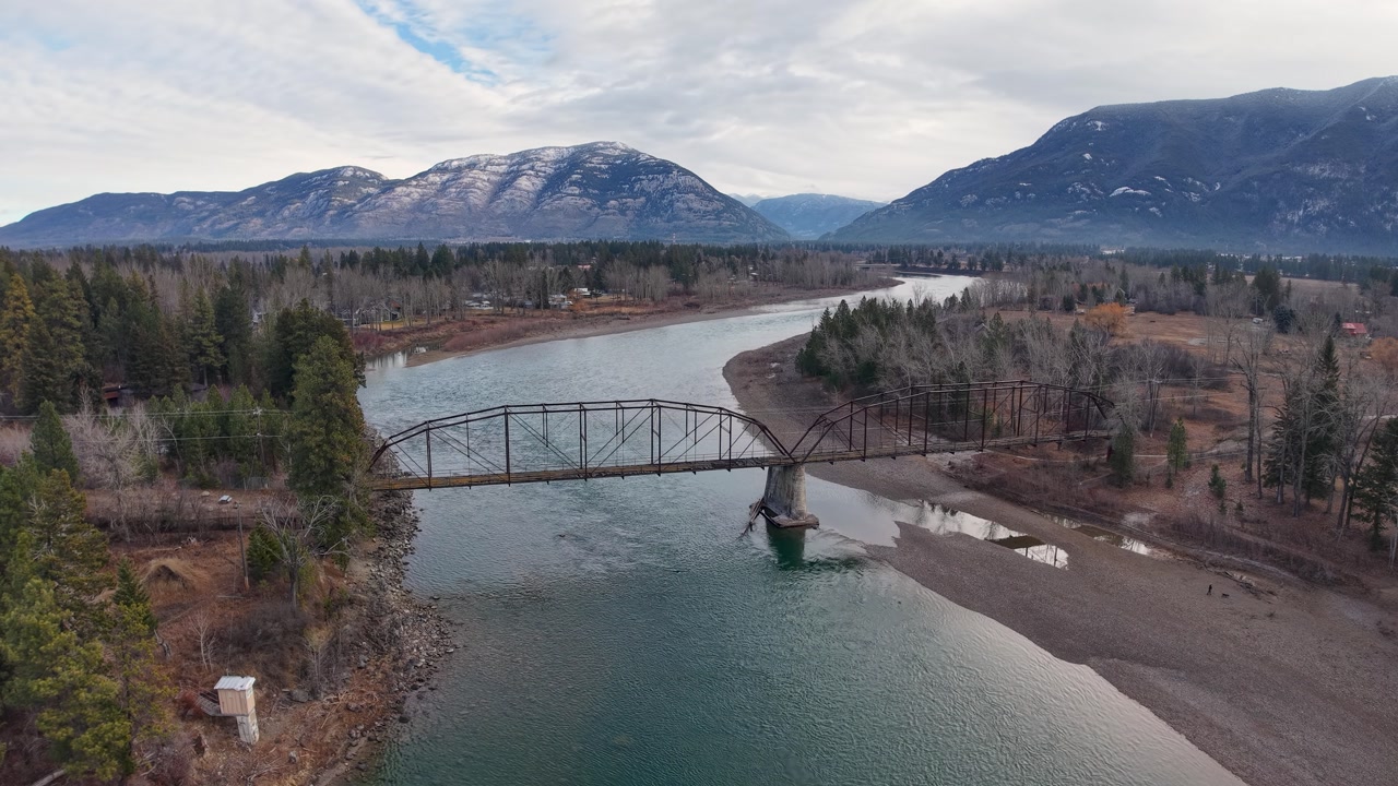 Aerial view of a metal truss bridge crossing a winding river with forested banks and snowcapped mountains in Montana.