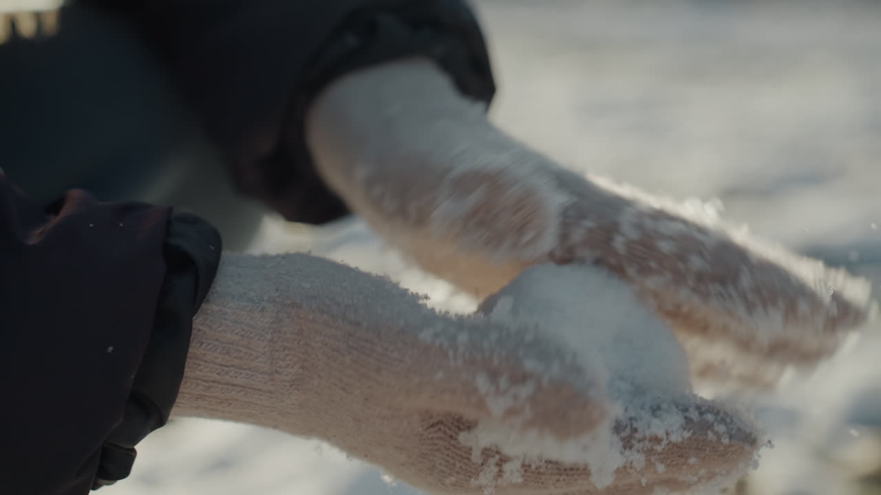Mãos enluvadas moldando uma pequena bola de neve, luvas de lã macias cobertas com neve fresca, campo nevado ao ar livre sob luz solar baixa, delicados cristais de gelo no tecido, close-up de gesto mostrando textura e movimento.