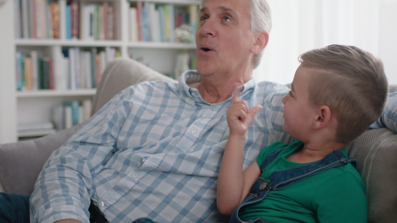 행복한 할아버지가 작은 소년에게 스마트폰을 사용하는 방법을 알려주고 호기심 많은 손자에게 현대 기술을 가르치고 지능적인 아이가 소파에 할아버지와 함께 앉아 휴대폰을 배우고 있습니다.