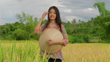 Mujer-Campesina-Vietnamita-Parada-En-Un-Campo-De-Arroz-Mirando-A-La-Cámara-Sonriendo-En-Cámara-Lenta-Sosteniendo-Un-Sombrero-De-Bambú-De-Arroz-Con-Ropa-Tradicional-De-Granja