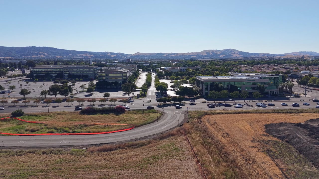 Un video drone mostra un'ampia vista aerea illuminata dal sole di moderni edifici per uffici, strade trafficate e una strada curva delimitata da campi aperti, con sullo sfondo colline distanti e un cielo azzurro brillante.