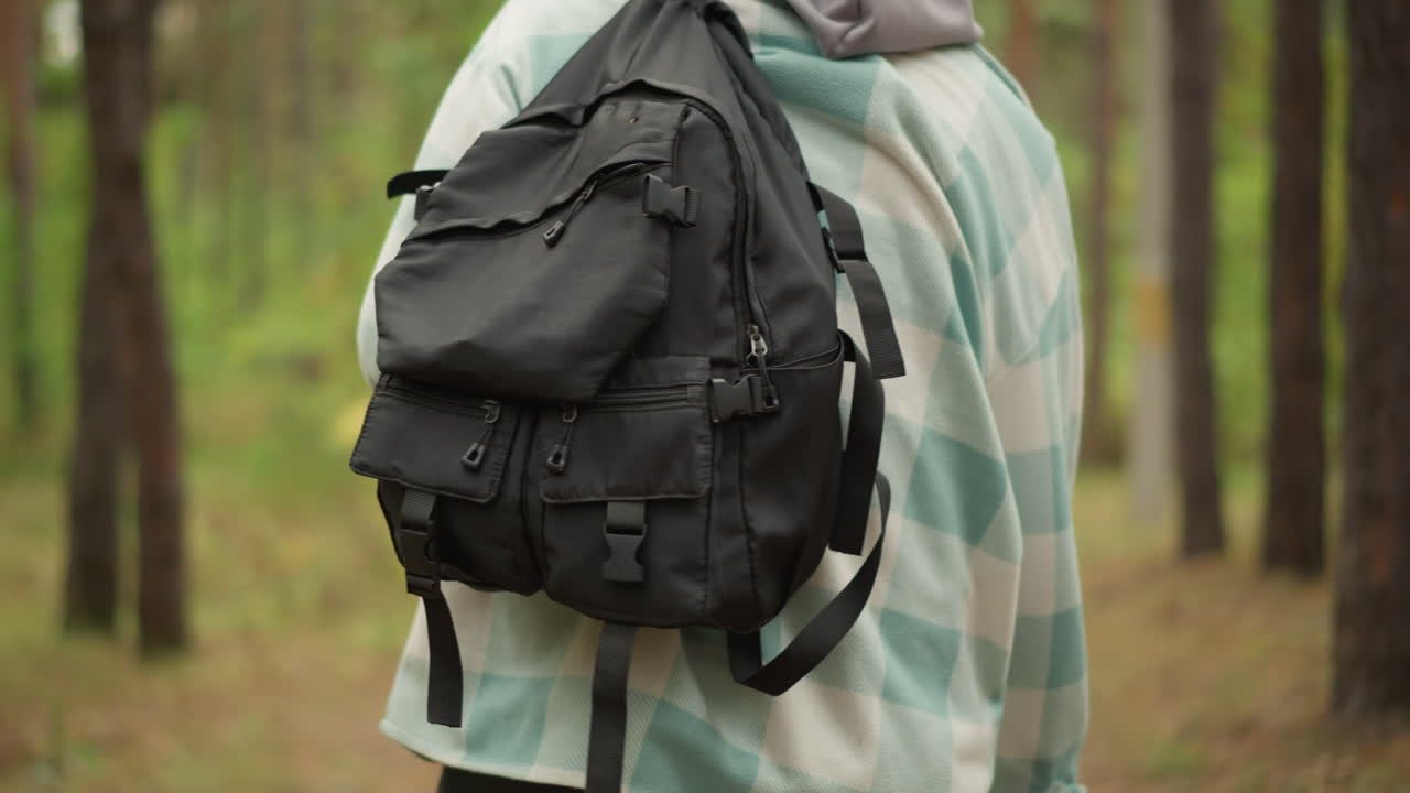 Reisender, der in der Nähe seinen robusten Rucksack inspiziert, Abenteurer, der in einer Waldumgebung seinen stabilen Sack prüft, Wanderer, der inmitten einer üppigen Baumumgebung und friedlichen Stimmung sorgfältig seinen abgenutzten Pack überprüft
