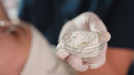 gloved hand of beauty therapist holds transparent glass cup filled with creamy white facial mask, ready for application, soft focus spa room, client in background enjoying facial treatment session