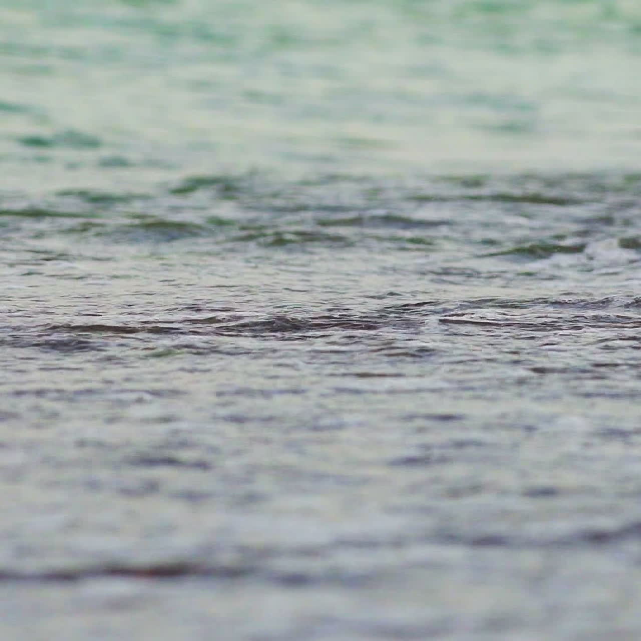 A turquoise sea waves cover the coast and take small grains of sand with it. Close-up. Blurred background.