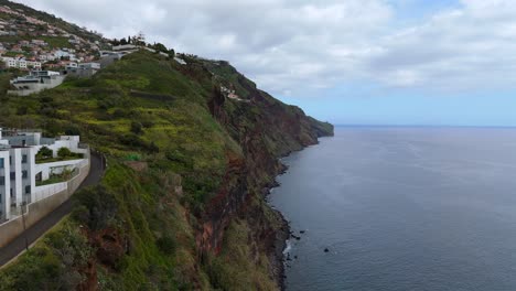 low drone flight along steep rocky coastline with modern architecture and green hillside rising above the atlantic ocean near câmara de lobos on madeira island