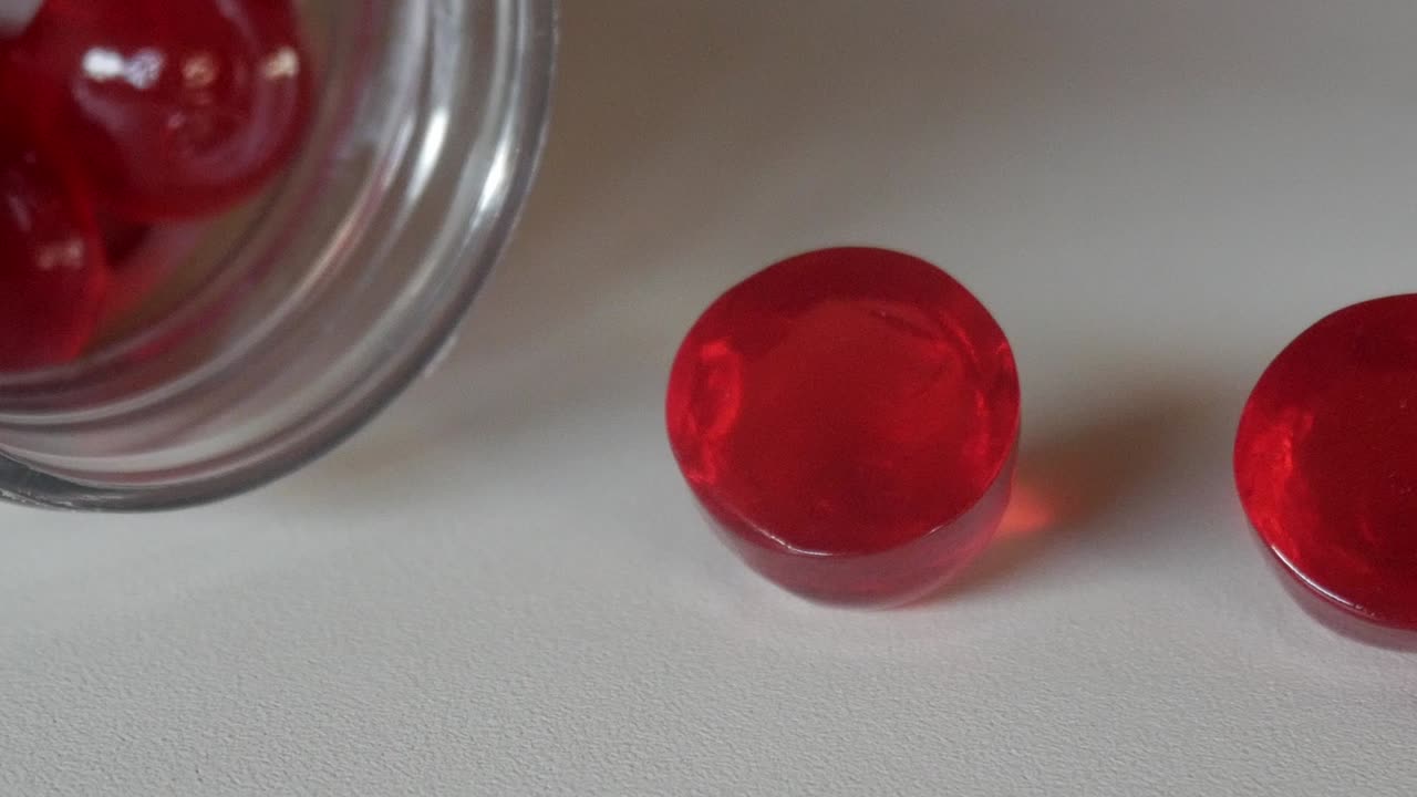 Close-up of red vitamin gummies spilling from a glass jar on a white surface under soft light