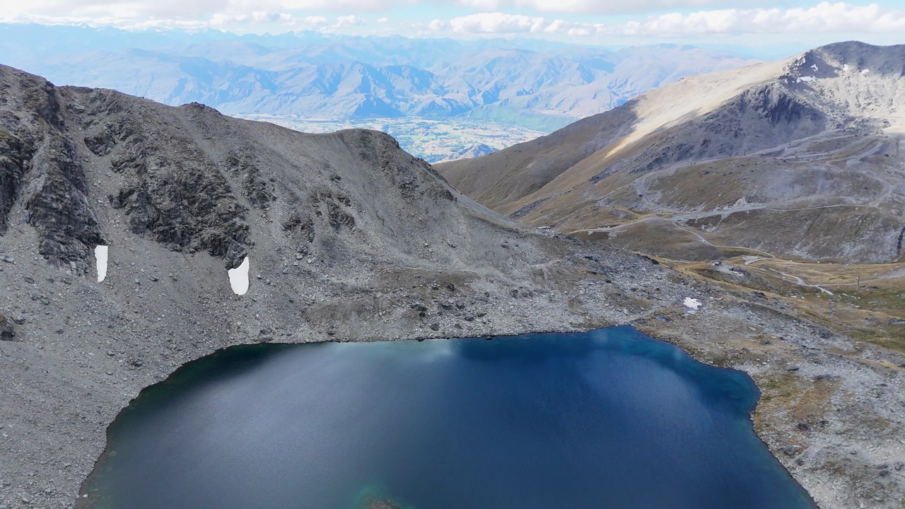 Aerial descends toward dark blue mountain lake on rugged alpine slope