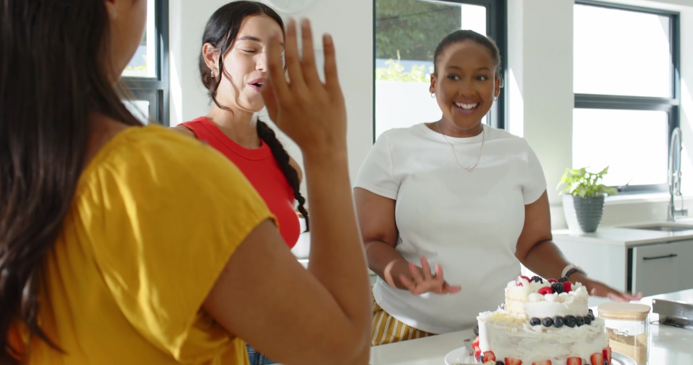 Diverse female friends spotting tiered cake, clapping, laughing on kitchen island, celebrating cake