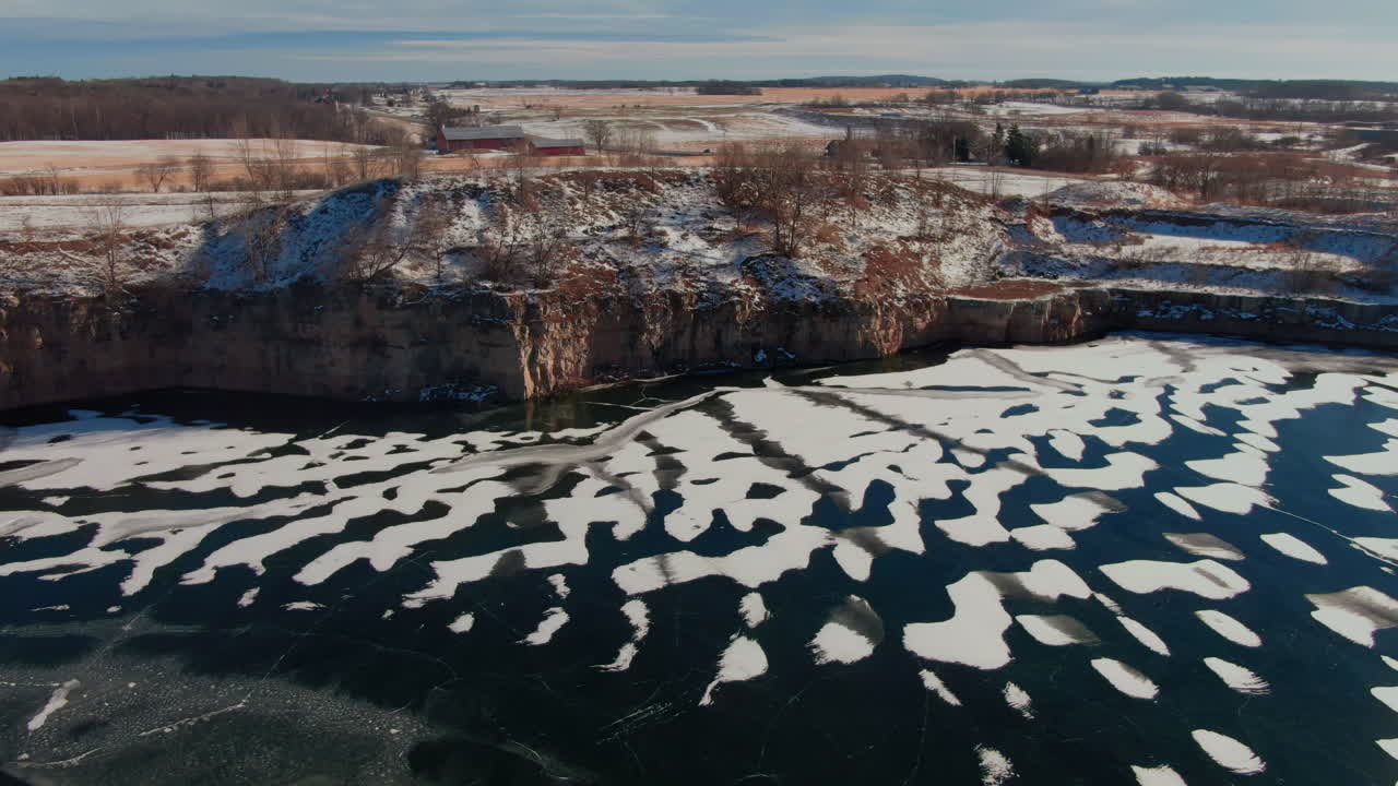 Ampia ripresa aerea di una cava ghiacciata con pareti rocciose ricoperte da un sottile strato di neve. Ghiaccio scuro e chiazze di neve creano un suggestivo paesaggio invernale