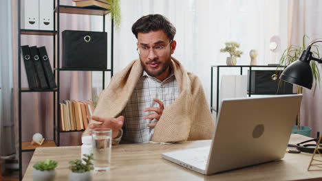 Sick-ill-Indian-businessman-wrapped-in-blanket-works-on-laptop-at-home-office-desk-takes-medicine