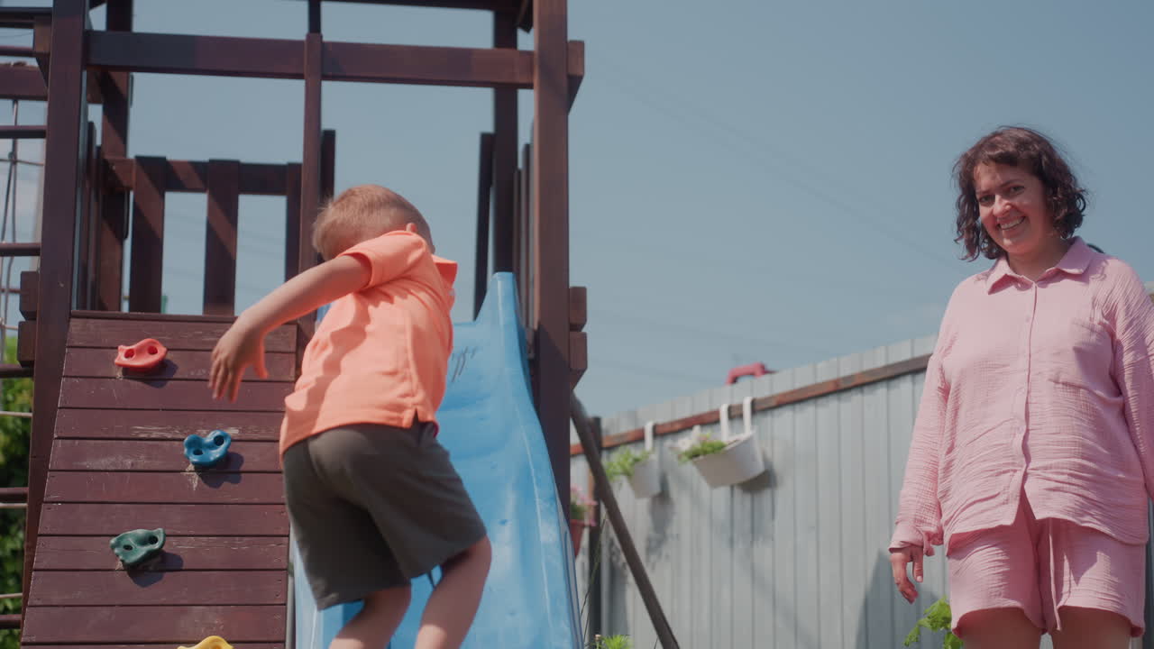 Boy At Blue Slide High Five With Smiling Mother Standing Nearby, Child Climbs Ladder And Returns For Celebratory Gesture, Sunny Yard, Playful Energy, Candid Family Interaction, Upbeat Confident Mood