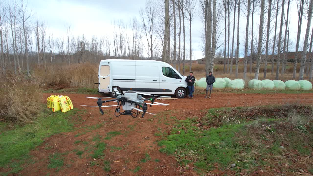 Veduta aerea di un drone agricolo che atterra su un sentiero fangoso vicino a un furgone e a due agricoltori che lo controllano.