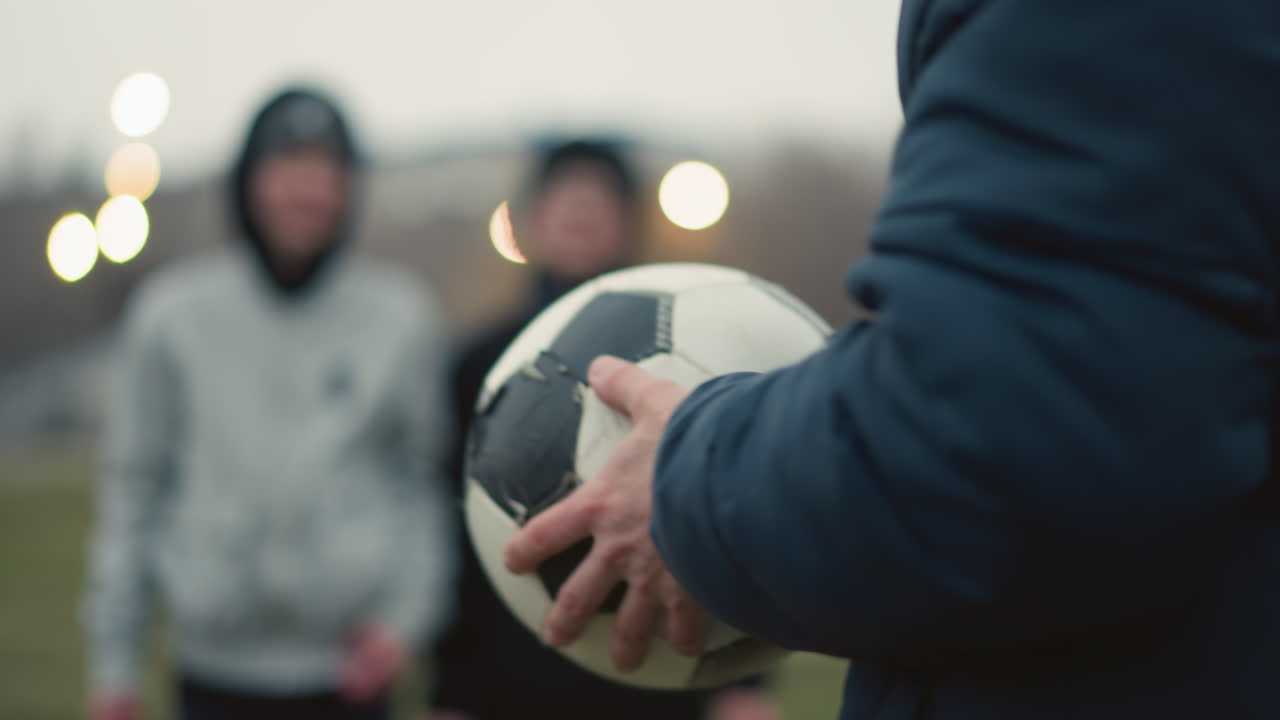 Close-up of a coach holding and turning a soccer ball in his hands, with a blurred view of two people standing in the background