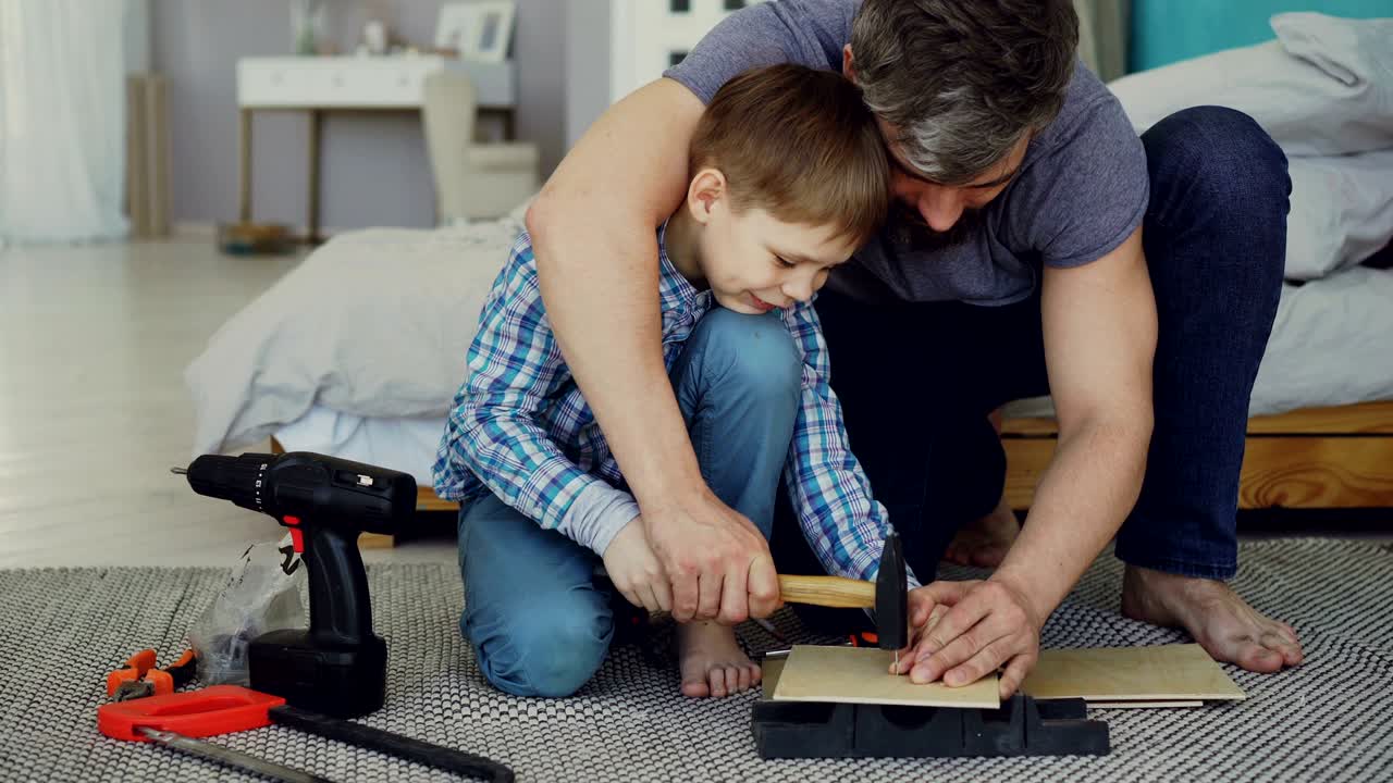 padre hombre barbudo está enseñando a su hijo cómo usar martillo clavar clavos en una pieza de madera juntos sentados en el suelo en casa. instrumentos, herramientas y muebles son visibles.