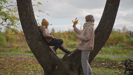 loving mother walks to daughter resting on tree in autumn park handing golden leaf and pecks daughter on cheek creating warm family moment amid vibrant fall foliage and serene nature scenery