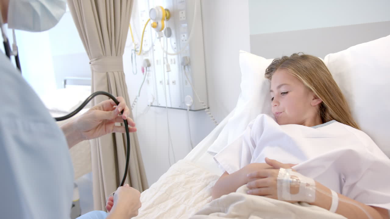 Caucasian female doctor in face mask using stethoscope on girl patient in hospital bed, slow motion