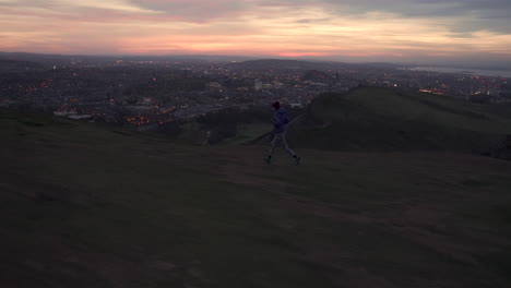 Toma-De-Seguimiento-Lateral-De-Una-Niña-Corriendo-En-La-Montaña-Arthurs-Seat-Por-La-Noche,-Al-Atardecer-Con-Las-Luces-De-La-Ciudad-De-Edimburgo-En-El-Fondo-Durante-La-Maravillosa-Luz-Del-Atardecer-Y-La-Hora-Azul