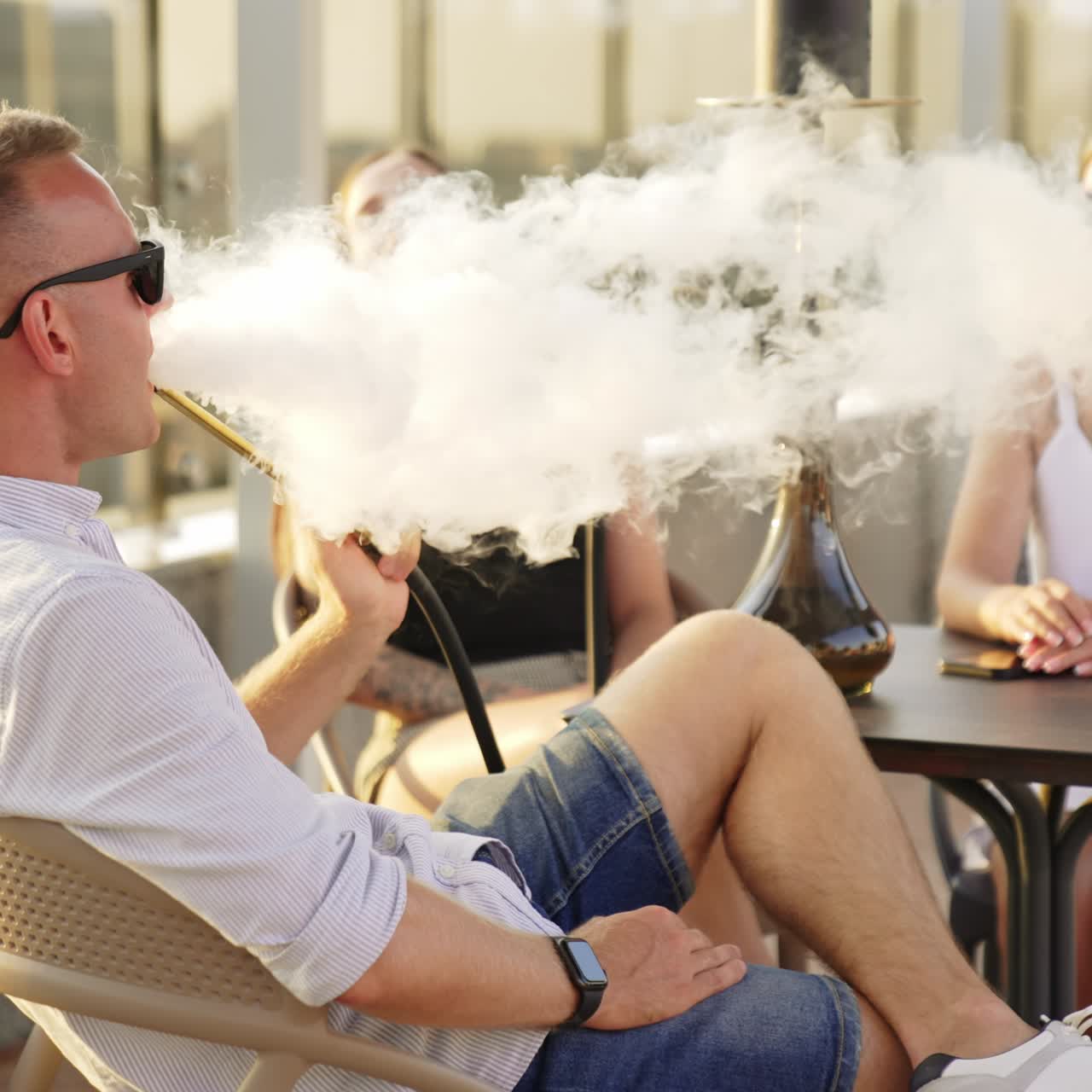 Company of happy relaxed people sitting outdoors at the table. Male smoking hookah and talking to female friends. Blurred backdrop
