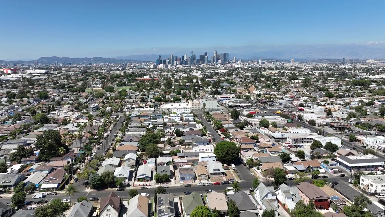 Riprese aeree con drone che mostrano lo skyline del centro di Los Angeles in lontananza oltre un denso quartiere residenziale in un luminoso e soleggiato pomeriggio estivo