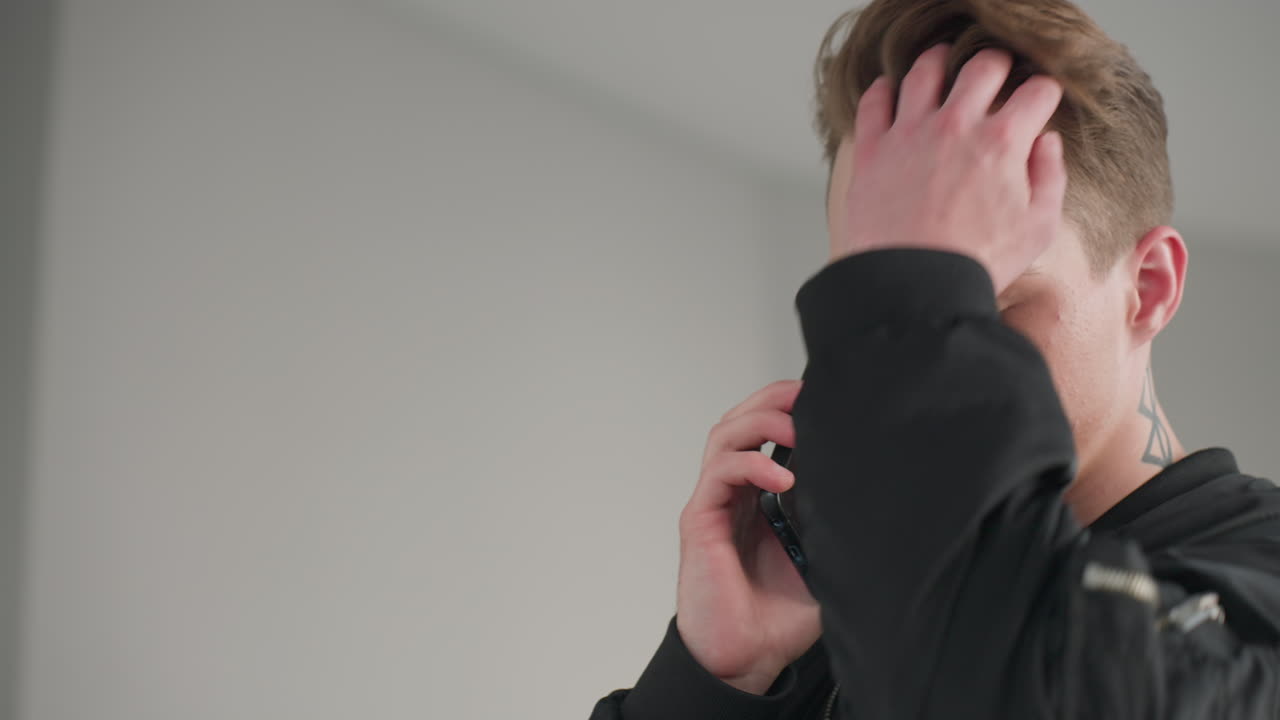 Teenager with geometric neck tattoo holds smartphone to ear speaking, uses free hand to push back hair while standing in bright minimalist room wearing black jacket