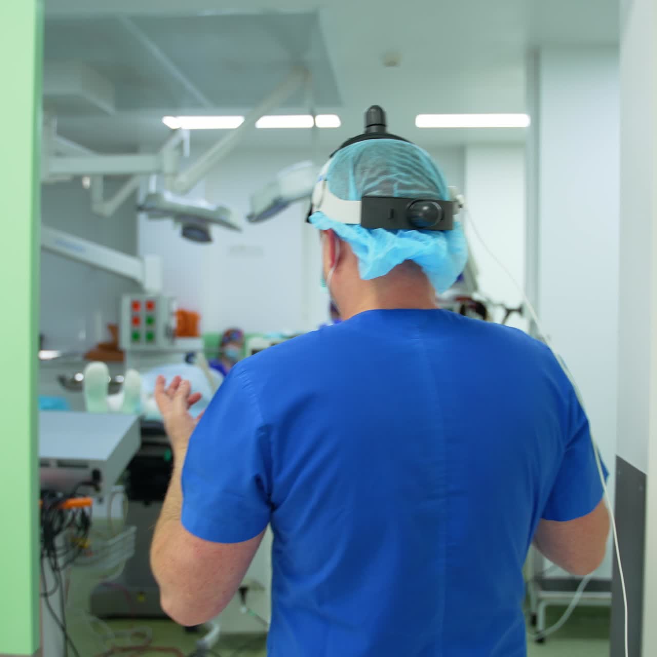Male doctor in uniform and device flashlight on his head heading to the surgery room. Specialist enters the operational theatre with prepared patient